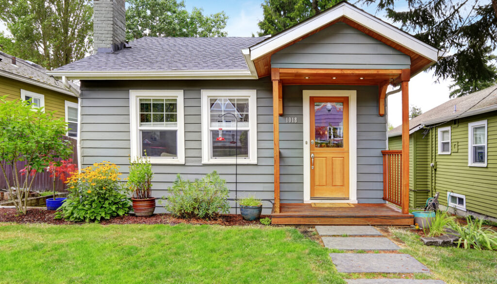 Small grey house with wooden deck. Front yard with flower bed and lawn